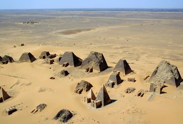 Seen from above, the pyramid field at Meroe reveals the true scale of this royal necropolis — dozens of steep-sided tombs clustered across the desert hills on the Nile's east bank, each once marking the resting place of a Kushite king, queen, or noble. The aerial vantage captures what ground-level views cannot: the sheer density of more than two hundred pyramids spread across a landscape that has remained largely unchanged for over two thousand years.