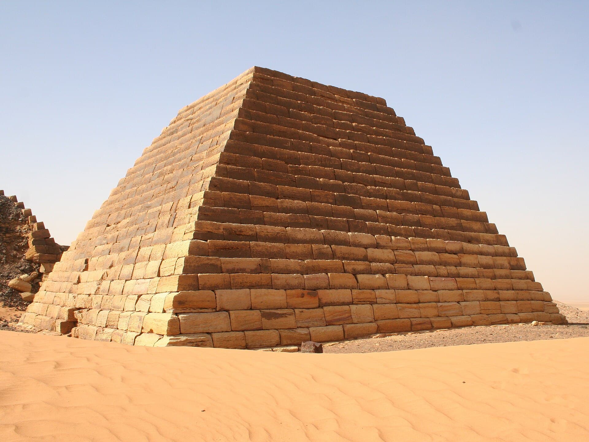 A single pyramid at Meroë seen up close, its sandstone courses laid with remarkable precision nearly two thousand years ago. Faint graffiti etched into the stone by 19th-century European visitors is still visible today — a reminder of the site's long and sometimes turbulent history of discovery and exploration.