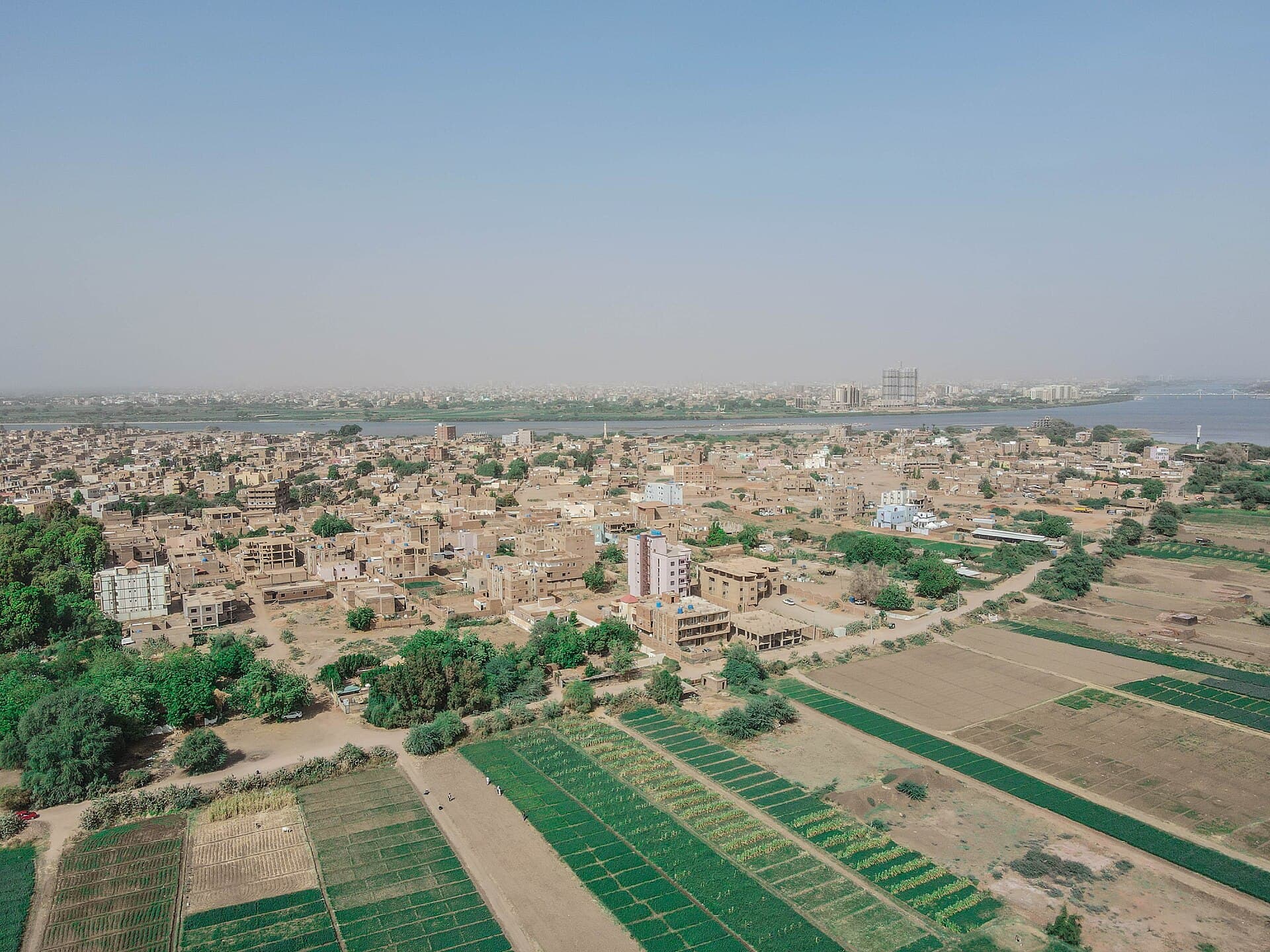 Seen from above, Tuti Island reveals its dual character: neat rows of vegetable fields stretching toward the Nile on one side, and a dense, growing neighbourhood of mudbrick and modern homes on the other — the capital's garden and one of its most distinctive communities, side by side.