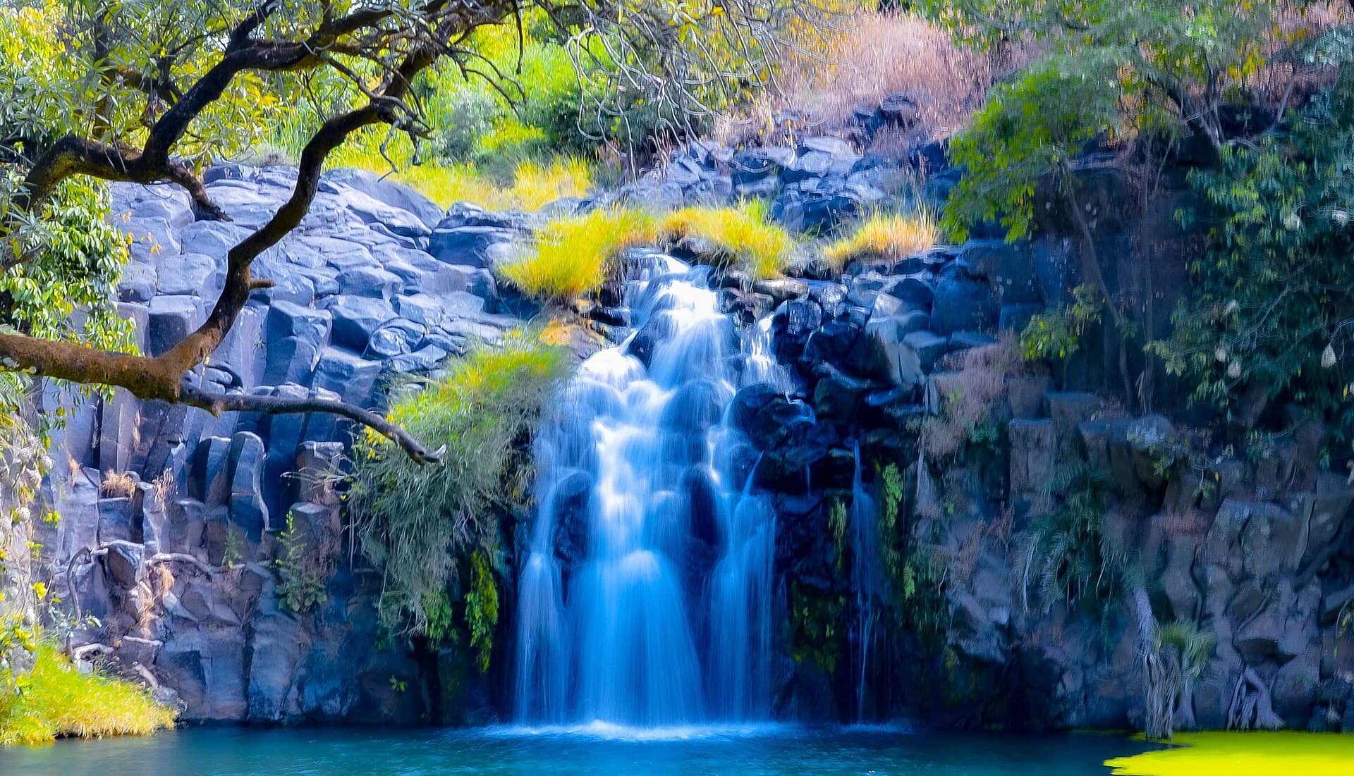 Water cascades over dark basalt columns on the slopes of Jebel Marra, feeding a clear turquoise pool below — a hidden waterfall in one of the volcanic highland's forested ravines.